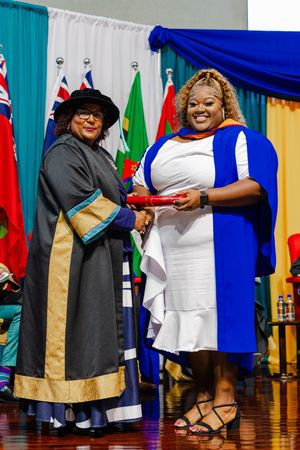 Two women in academic regalia exchanging a diploma at a graduation ceremony with international flags in the background.