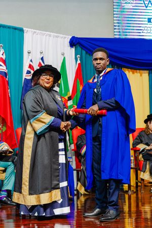Graduate in blue robe receives award from official in ceremonial dress at graduation event.