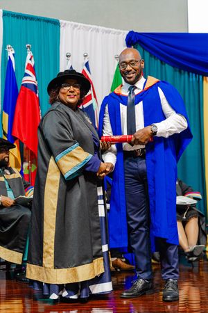 Two graduates in academic regalia shake hands during a university graduation ceremony with flags in the background.