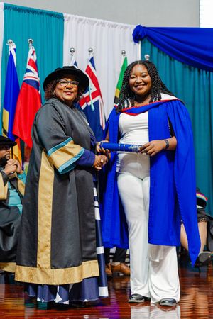 Two women in graduation regalia smiling while exchanging a diploma on stage at a graduation ceremony.