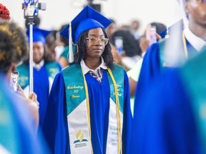 Students in blue and green graduation gowns and caps walk in a procession at a graduation ceremony.