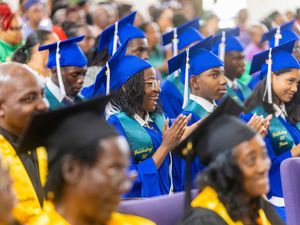 Graduates in blue caps and gowns celebrate at a graduation ceremony, with a smiling woman clapping in the foreground.