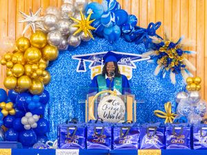 Graduate in blue cap and gown stands at podium surrounded by gold and blue balloon arch and grad gift bags.