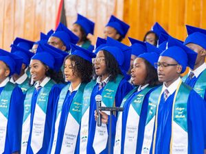 Students in blue graduation caps and gowns with green stoles celebrate at a graduation ceremony.