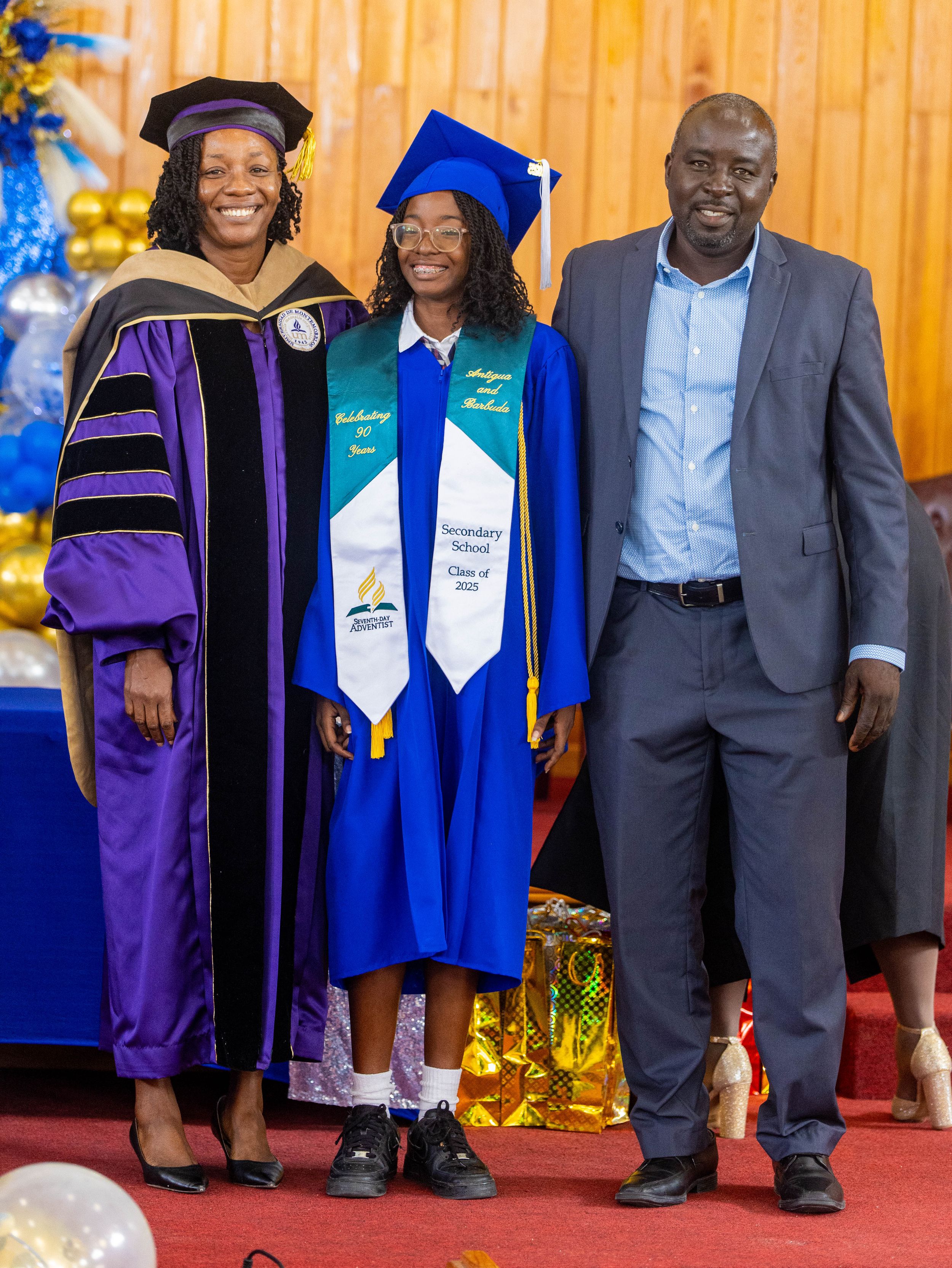 Graduate in blue cap and gown poses with two proud family members at a secondary school graduation ceremony.