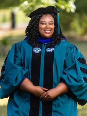 Smiling graduate in teal doctoral regalia with black cap and blue hood poses outdoors for graduation portrait.