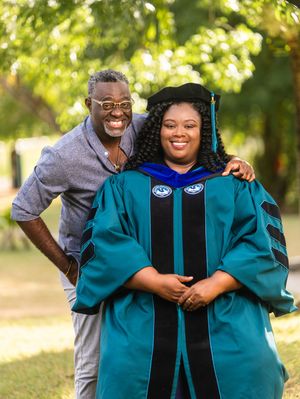 A proud graduate in teal doctoral regalia poses with a smiling man outdoors during a graduation celebration.