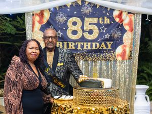 Couple posing together at an elegant 85th birthday celebration with gold and black decorations and a festive cake.
