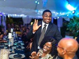 Smiling man in a suit waving at guests during a formal evening birthday party with elegant table settings.