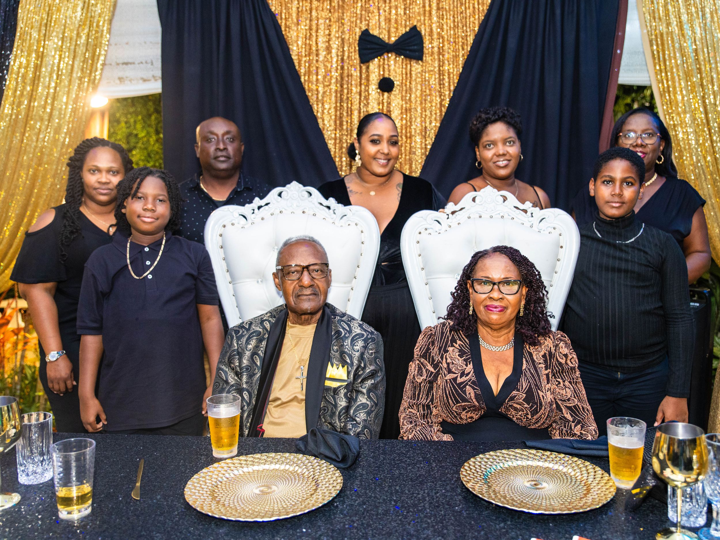 Family group photo at elegant black and gold formal event, with elderly couple seated in ornate white chairs at decorated table.