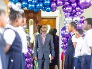 Man in gray suit walks through balloon arch of blue, purple, silver balloons, flanked by students in school uniforms.