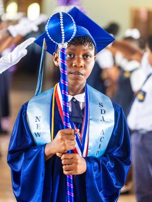 Student in blue cap and gown holds ceremonial mace at New SDA Academy graduation.