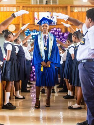 Graduate in blue regalia walks through honor guard arch at SDA Academy commencement ceremony.