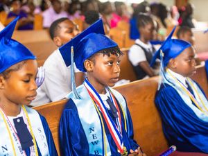 Young students in blue graduation caps and gowns seated in church pews during a graduation ceremony.
