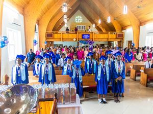 Class of 2025 graduates in blue caps and gowns stand in decorated church for graduation ceremony.