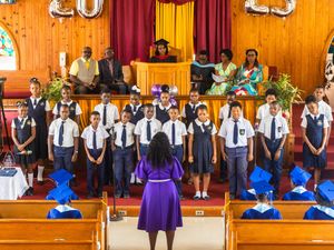 School choir performs in church, students in uniforms directed by teacher in purple dress during graduation ceremony.