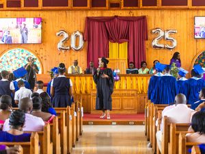A graduation ceremony in a decorated church with '2025' balloons, graduates in blue robes, and a speaker at the podium.