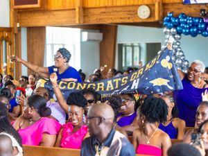 Joyful crowd celebrates graduation in a church, holding a Congrats Grad banner, dressed in blue and purple attire.