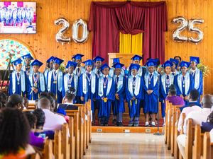 graduates in blue caps and gowns stand on stage under 2025 balloons at a school graduation ceremony.