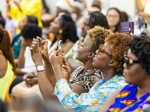 Audience of Black women at an event, taking photos with smartphones, wearing colorful attire.