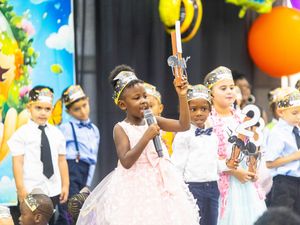 A young girl in a pink gown holds a microphone on stage during a colorful kindergarten graduation ceremony.