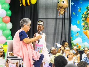 Young girl in tiara receives microphone at colorful bee-themed children's event with balloon decorations.