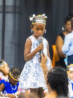Young girl in blue floral dress and decorative crown speaks into microphone at cultural event.