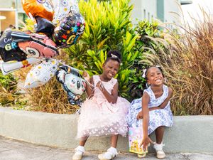 Two young girls in pretty dresses smile while sitting outdoors with colorful character balloons.