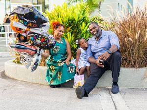 Family celebrating graduation with balloons outdoors, woman in teal dress, man in blue shirt, child between them.