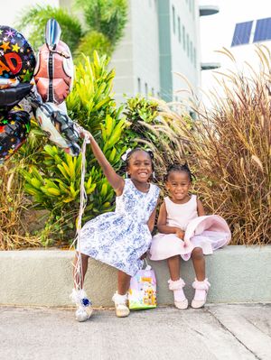 Two young girls in dresses holding graduation balloons while sitting outdoors on a sunny day.