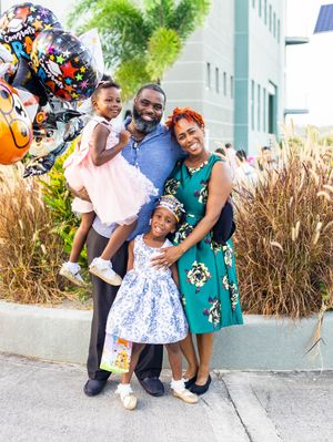 Family of four posing outdoors with graduation balloons, parents in festive attire with two young daughters.