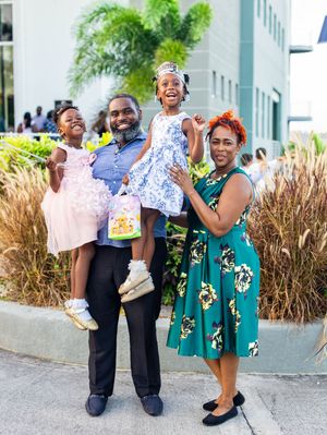 Happy family of four posing outdoors, parents holding two young daughters in colorful dresses, smiling joyfully.