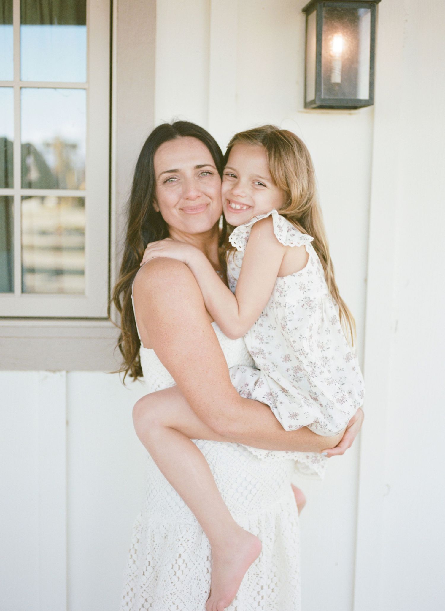 A smiling mother in a white lace dress holds her young daughter in a floral outfit outdoors near a lantern.