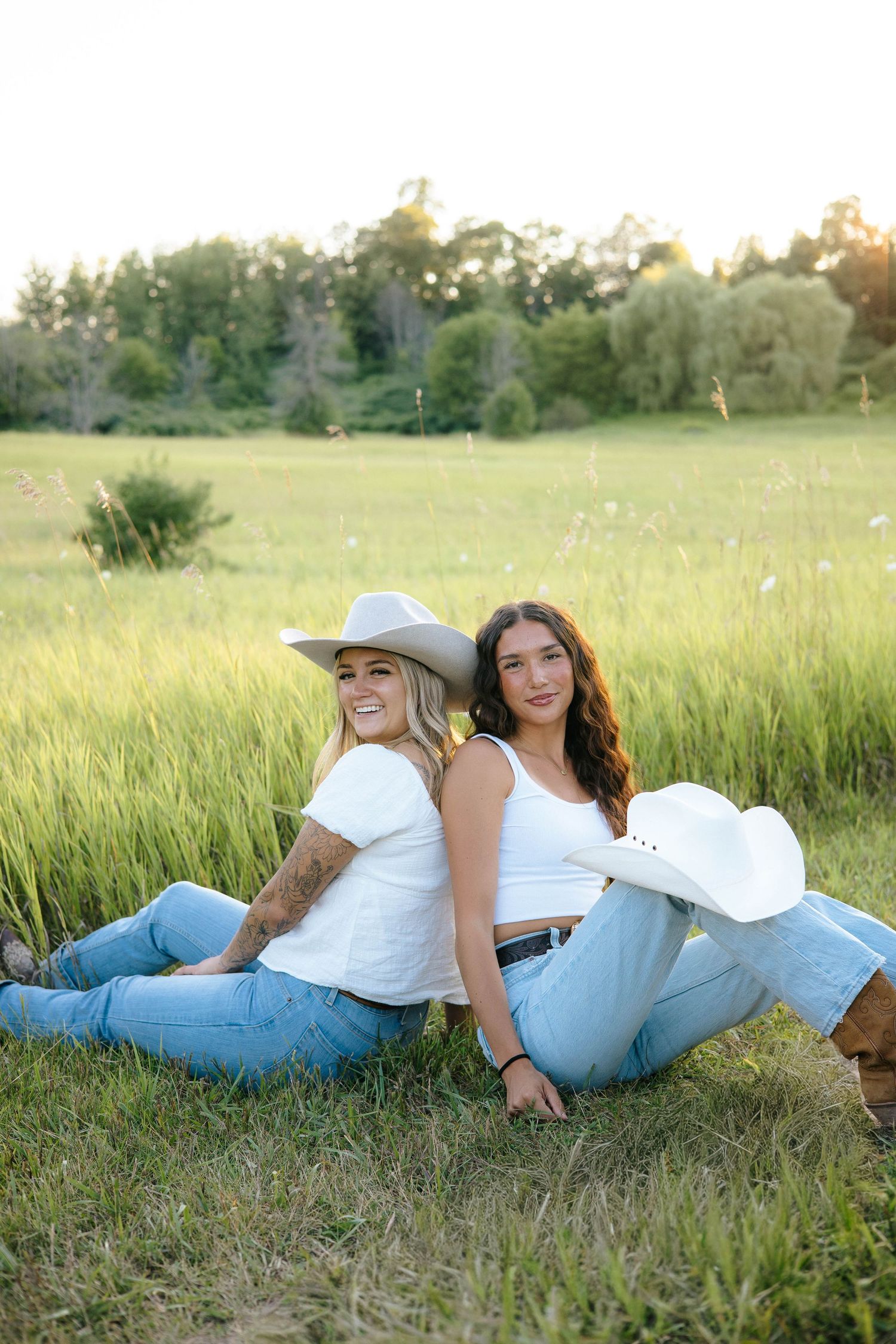 Two young women in white tops and jeans sit back-to-back in a green meadow, one wearing a cowboy hat, smiling at sunset.