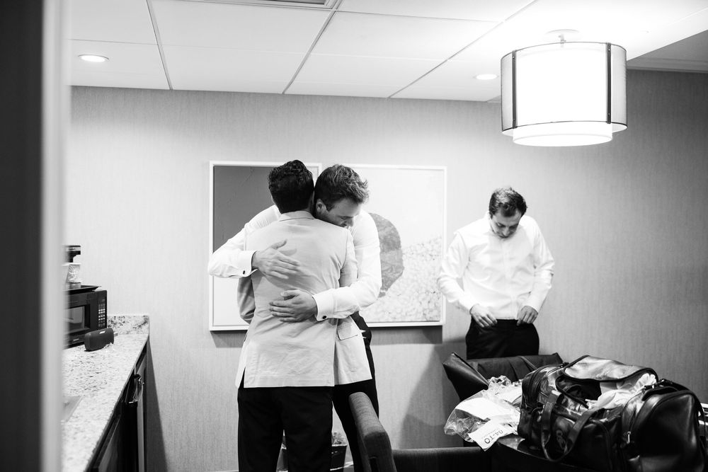 Black and white photo of groomsmen helping each other dress in a hotel room before a wedding ceremony.