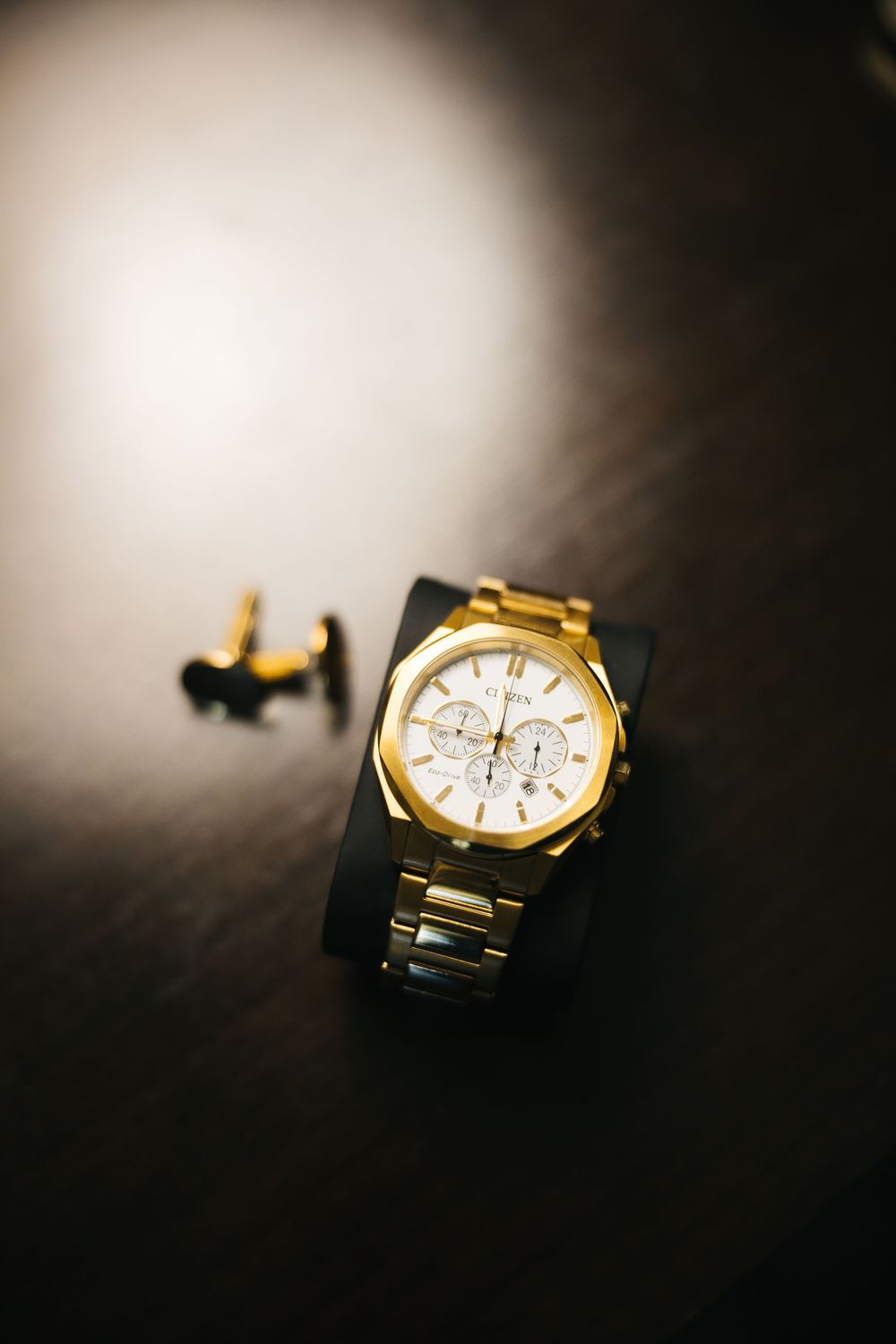Gold chronograph watch with white dial on dark surface beside gold cufflinks, elegant luxury timepiece accessory.