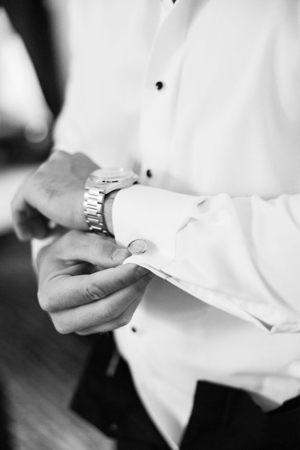 Groom fastening cufflinks on white dress shirt while wearing a silver watch, black and white wedding photo.