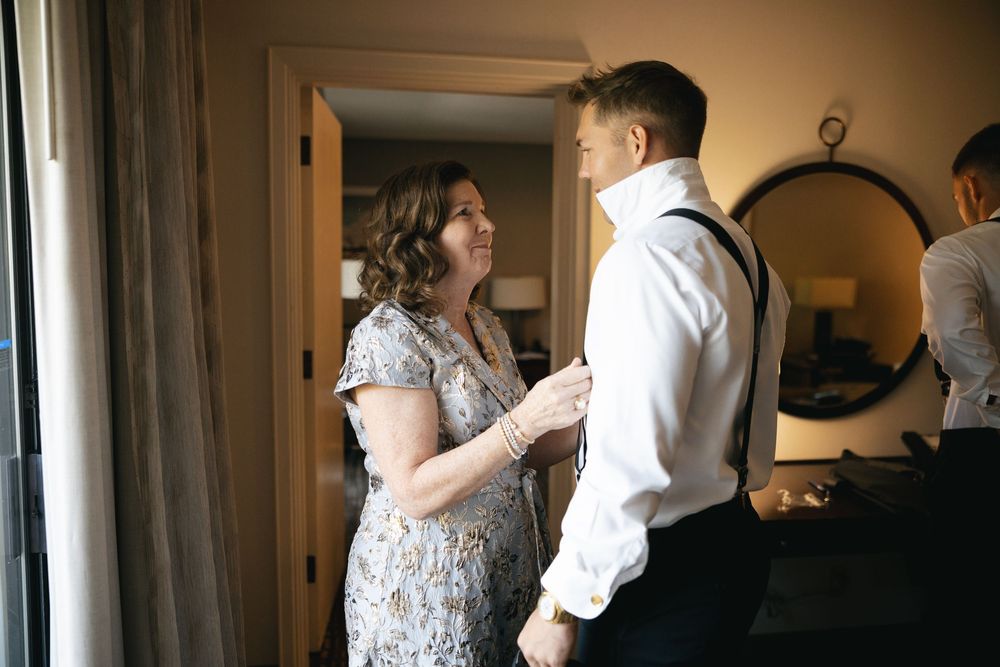 A woman in a floral dress helps a groom in suspenders button his shirt before a wedding ceremony.
