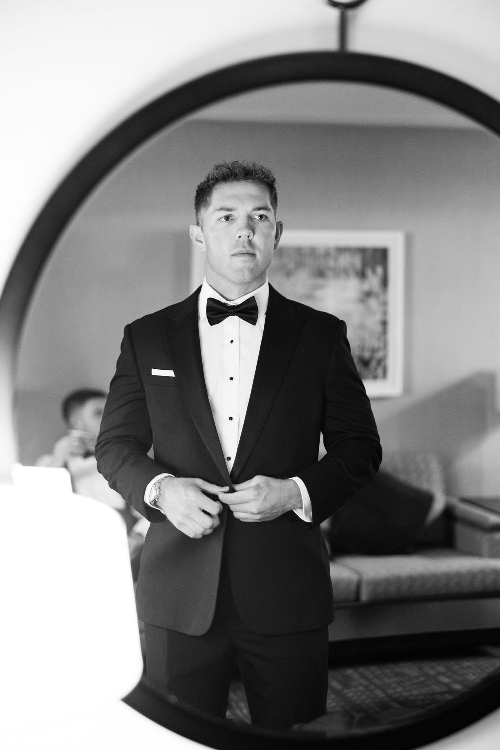 Groom in black tuxedo and bow tie buttoning jacket in hotel room, elegant black and white portrait.