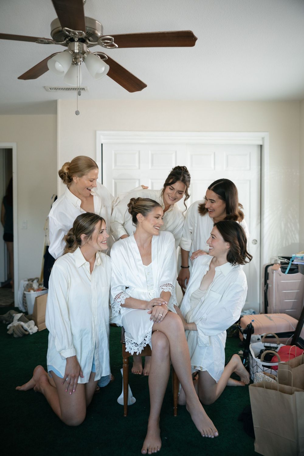 Bridesmaids in white robes laughing together while getting ready for a wedding in a cozy room.