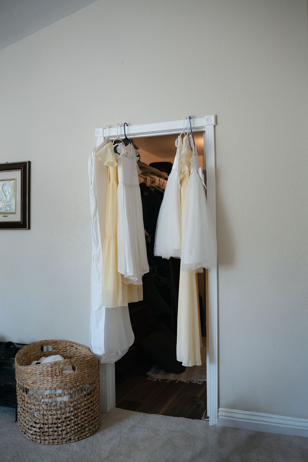 White and cream bridesmaid dresses hanging on a closet door next to a wicker basket in a neutral bedroom.