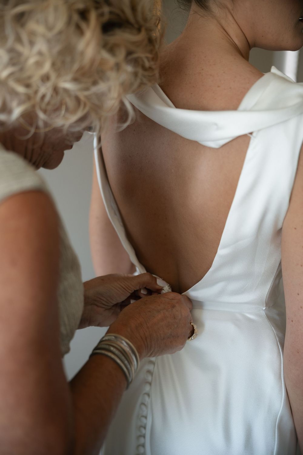 Woman in white open-back wedding dress being helped with buttons by blonde bridesmaid during bridal preparations.