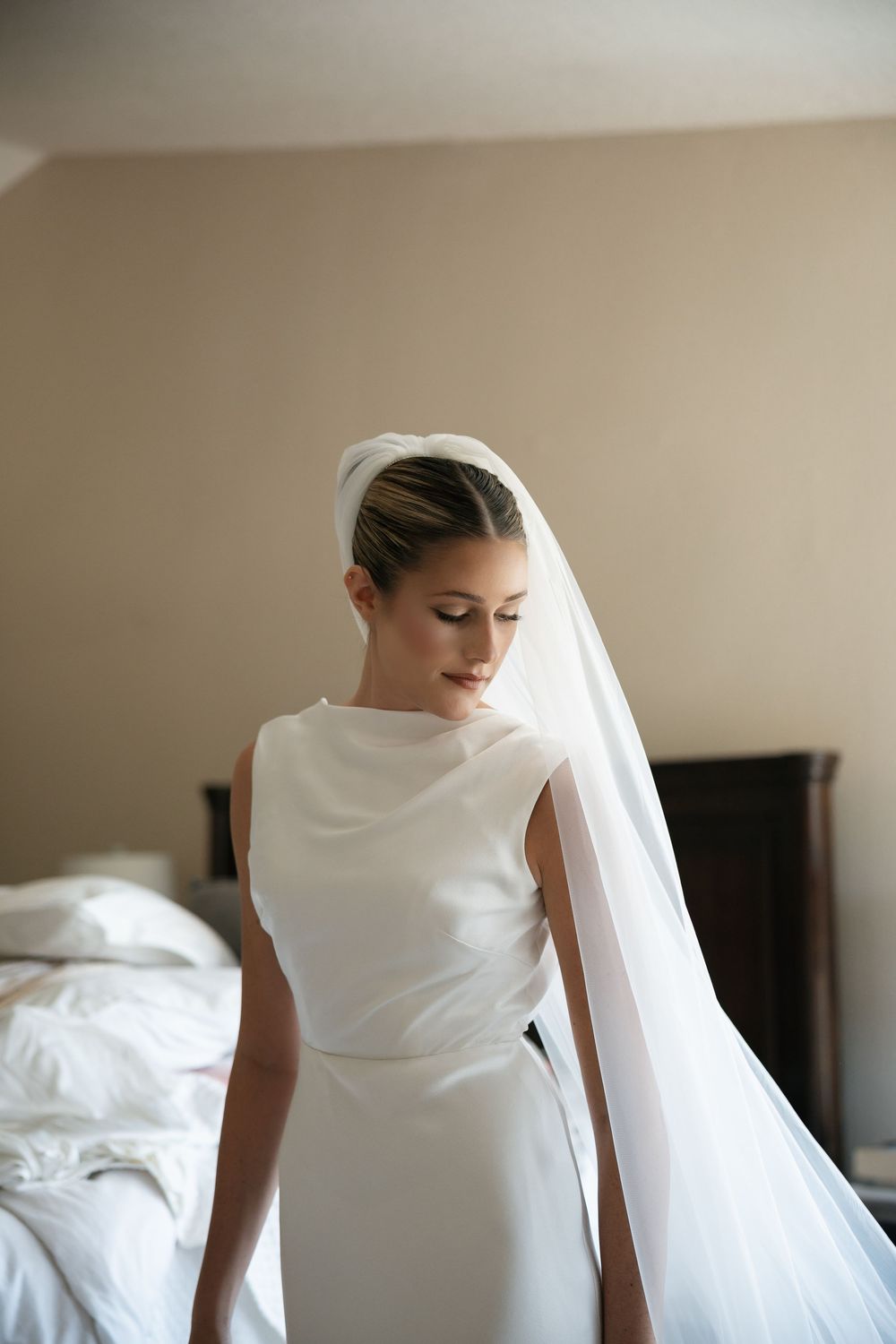 Elegant bride in sleeveless white satin gown with cathedral veil and updo hairstyle, looking down in a hotel room.