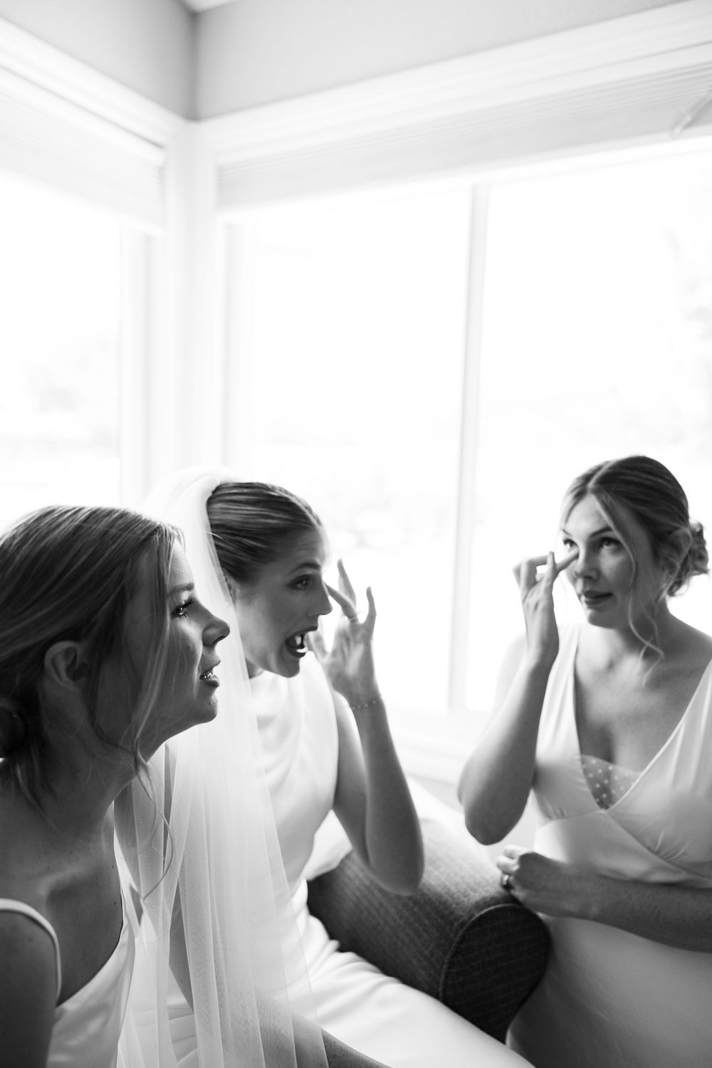 Bride and bridesmaids share joyful moment while getting ready near bright window in black and white.