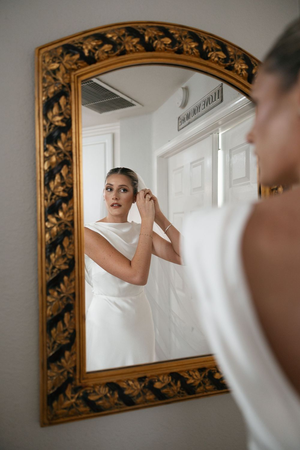 Bride in white gown adjusting her hair while looking into an ornate gold-framed mirror in a bright room.