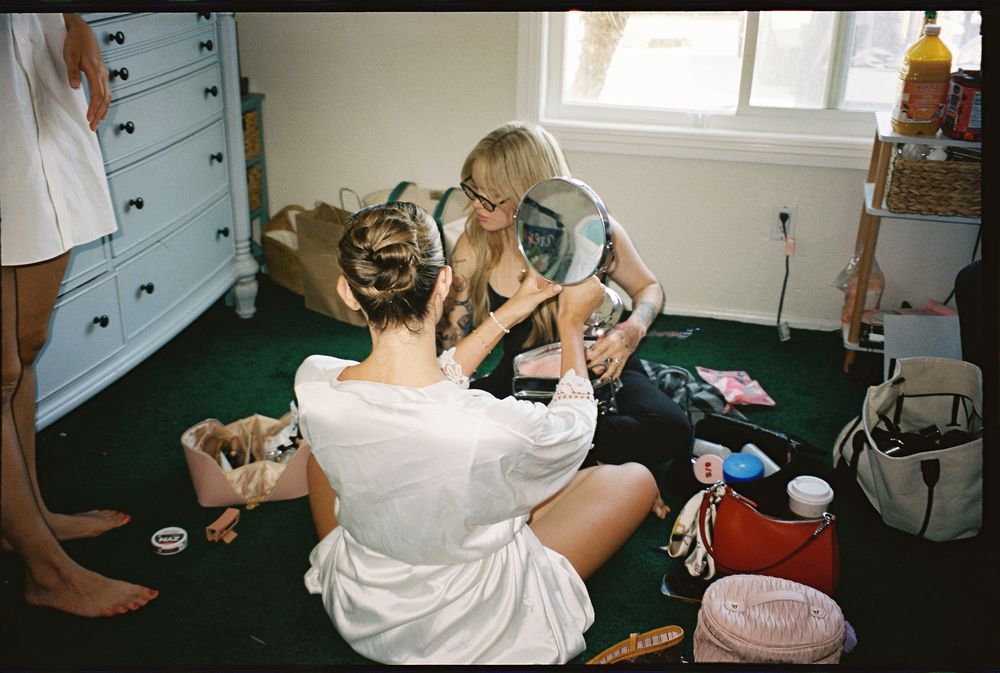 Two women sit on a carpeted floor getting ready, with a mirror, dresser, and beauty products surrounding them.