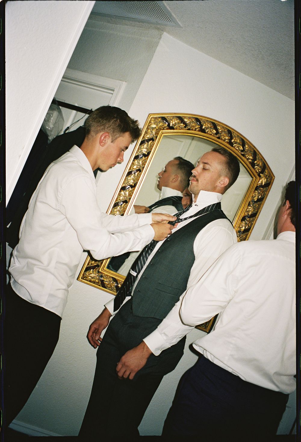 Groomsmen helping each other with ties before a wedding ceremony indoors near a gold-framed mirror.