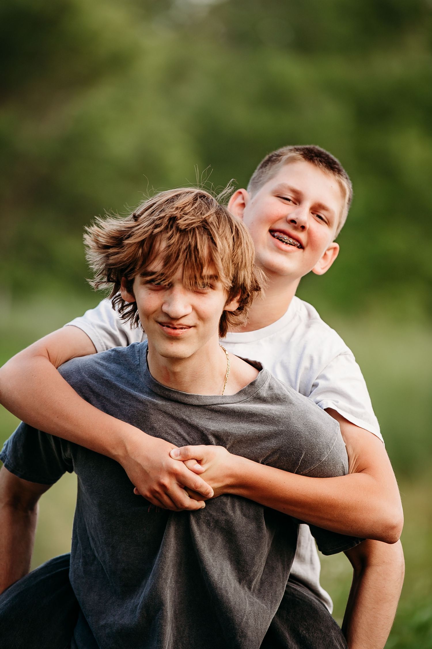Two teenage brothers laughing and sharing a piggyback ride outdoors in green nature.