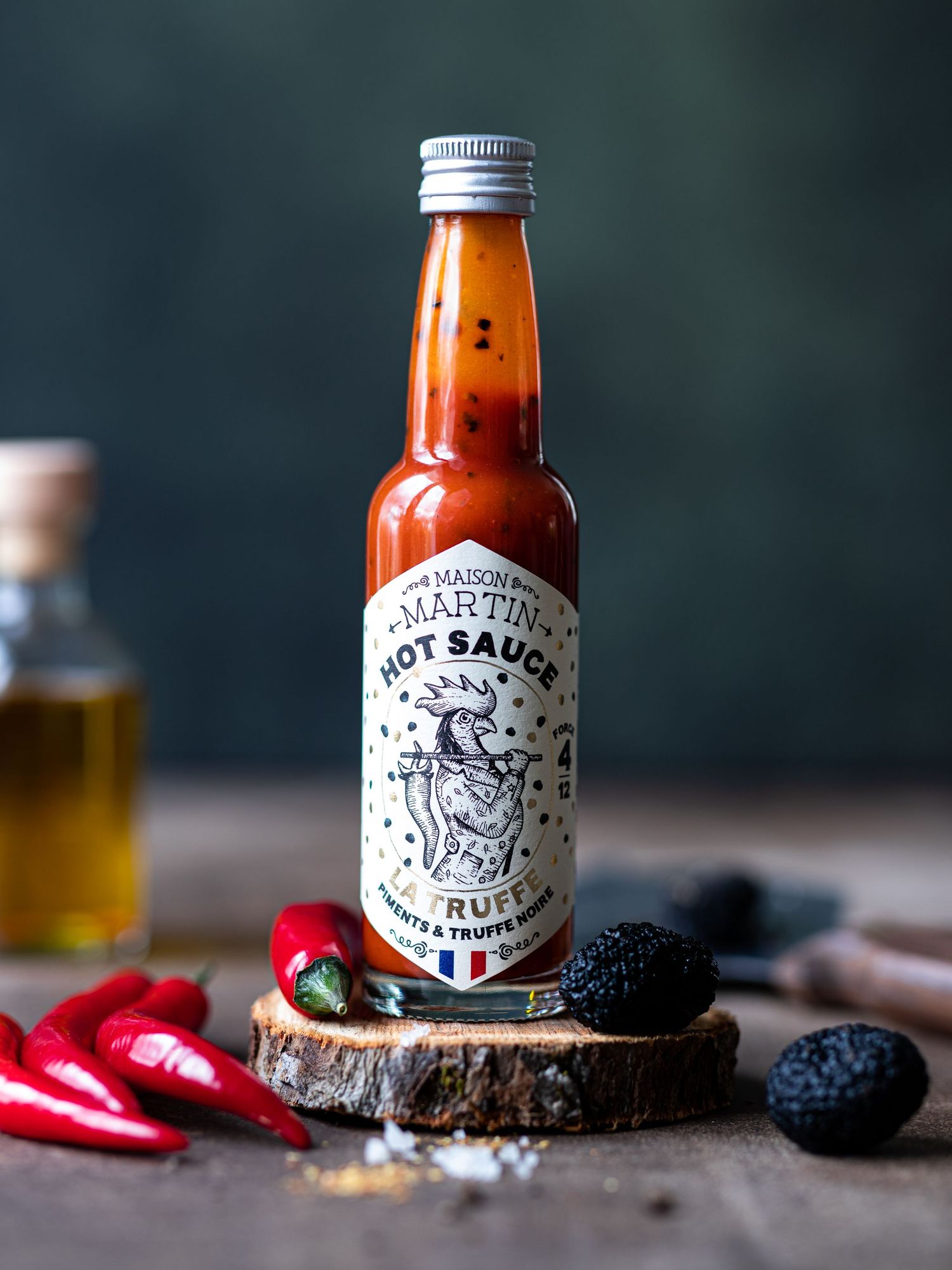 Maison Martin hot sauce bottle with truffle and chili peppers on a wooden slice, surrounded by fresh red chilies and black truffles.