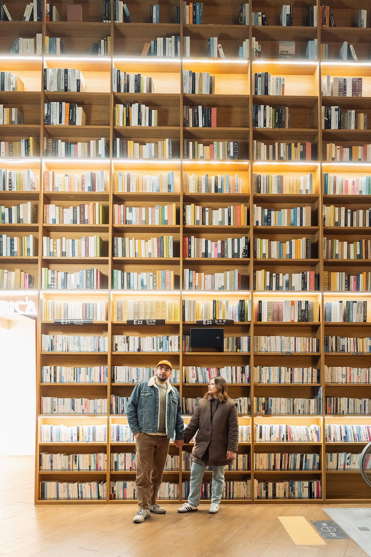Two people stand in front of a large floor-to-ceiling wooden bookshelf filled with books in a modern library.